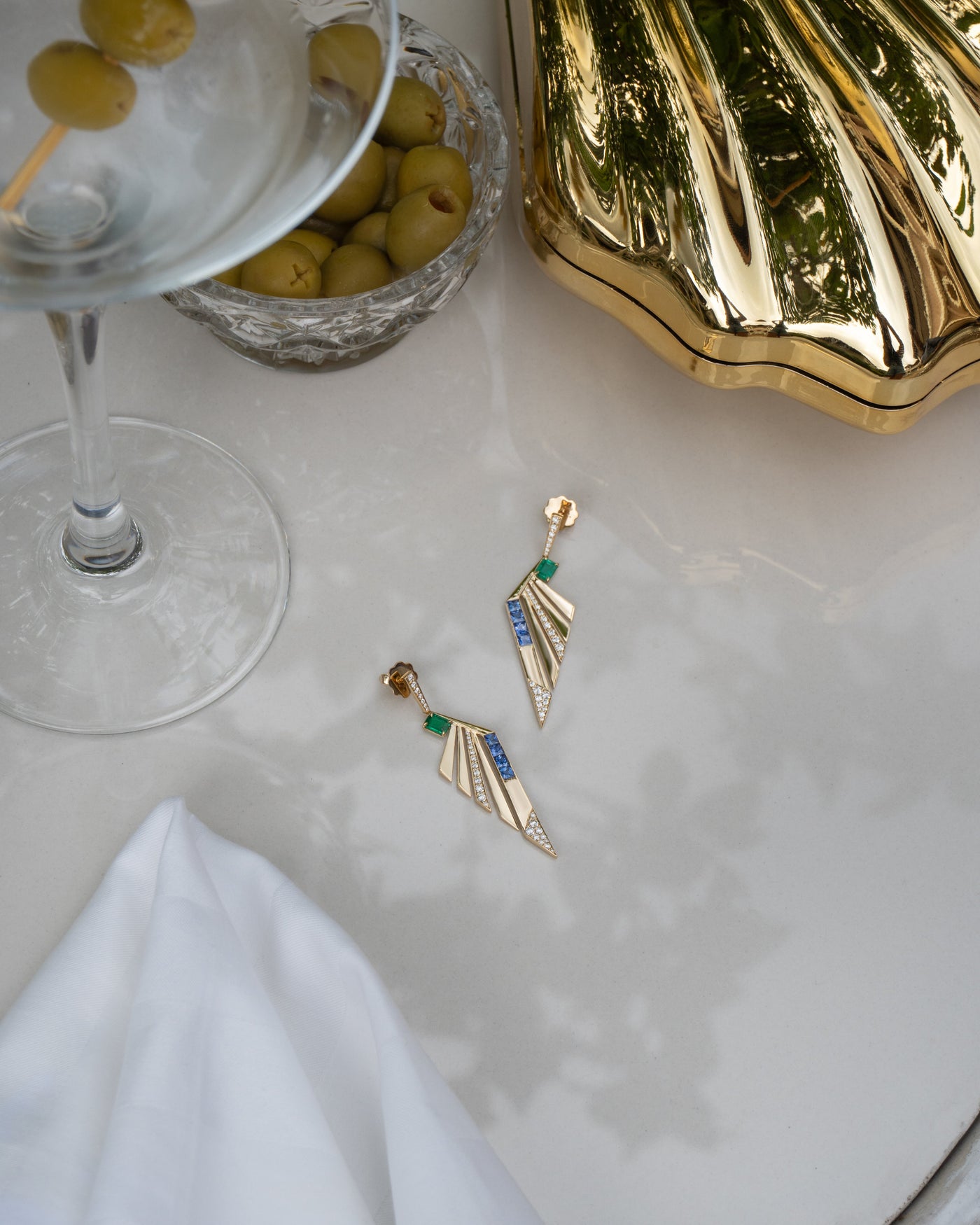 Gold earrings with gemstones on a white surface next to a cocktail glass with olives.