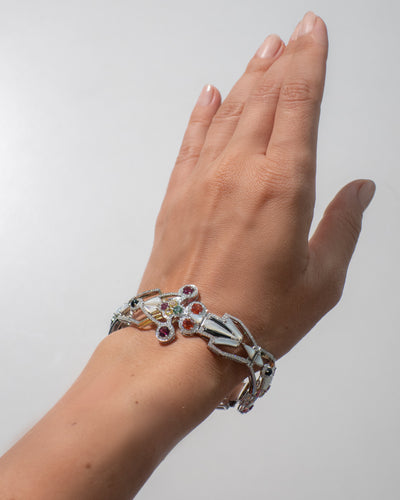 Hand wearing a gold bracelet with colorful stones against a plain background